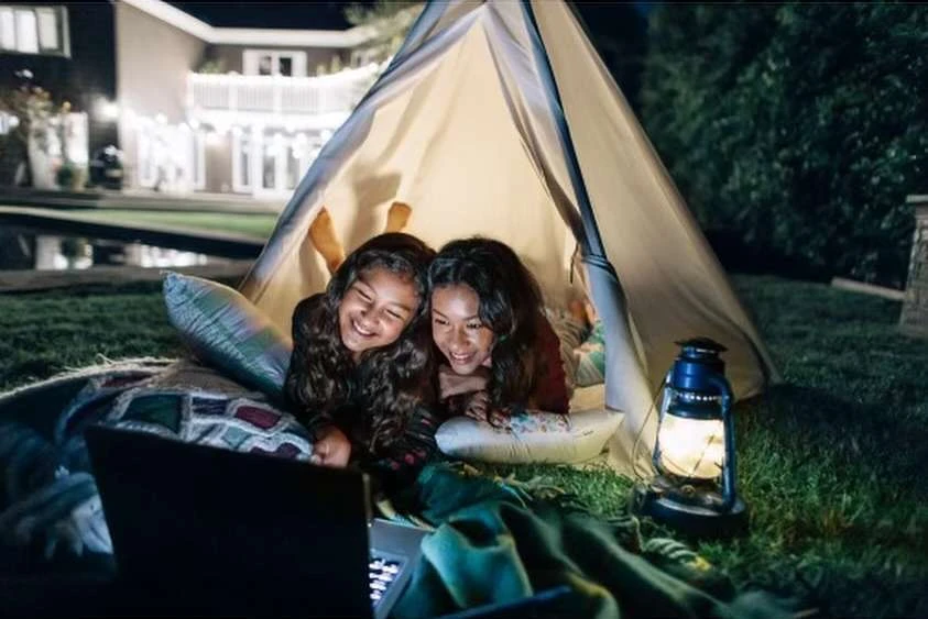Two smiling girls using a laptop inside a tent at night with a lantern. Outdoor sleepover with backyard, home, and trees visible.