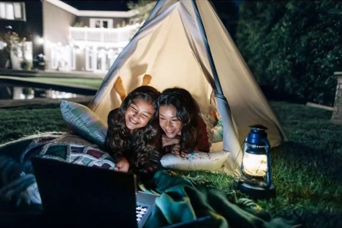 Two smiling girls using a laptop inside a tent at night with a lantern. Outdoor sleepover with backyard, home, and trees visible.