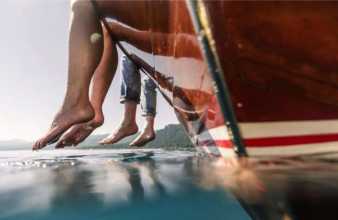 Two people sit on a boat with their legs dangling over crystal clear water. The background shows a forested shoreline.