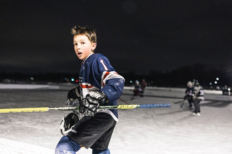 Young boy in hockey jersey on ice rink, playing under the night sky, holding hockey stick and helmet during a game.