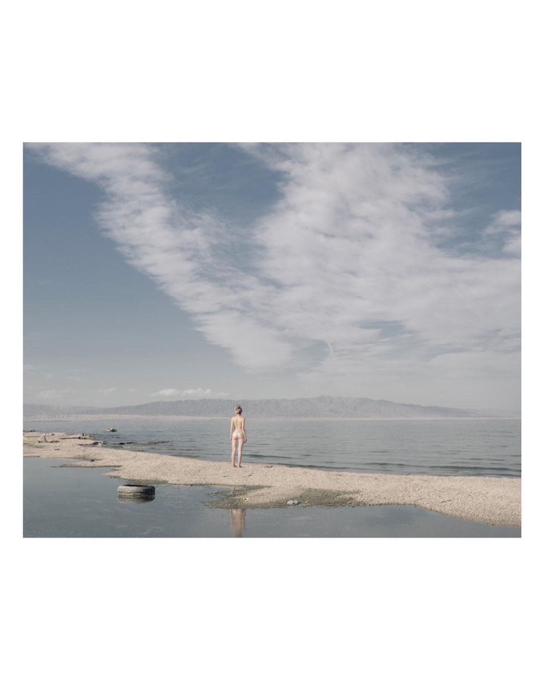 Woman in swimwear standing on a sandbar in a lake, mountains in the distance, blue sky with clouds.