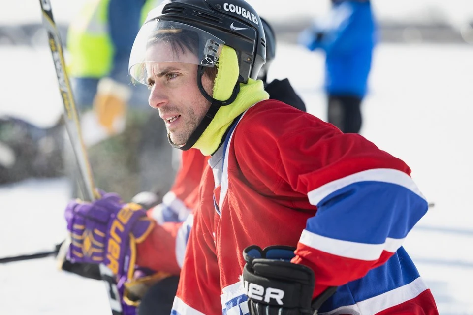 Adult hockey player wearing protective helmet and red, white, and blue jersey on outdoor ice rink with hockey stick and other players.