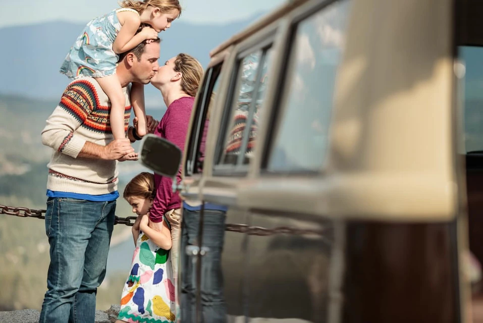 Family of four: father carries daughter on shoulders, mother kisses father, and another daughter stands near an older van outdoors.