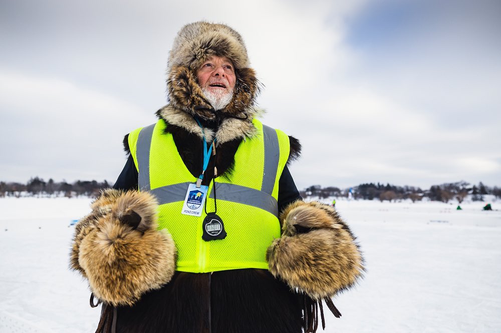 Older man with a fur hat and large fur mittens stands in a snowy landscape wearing a yellow safety vest and an ID badge.