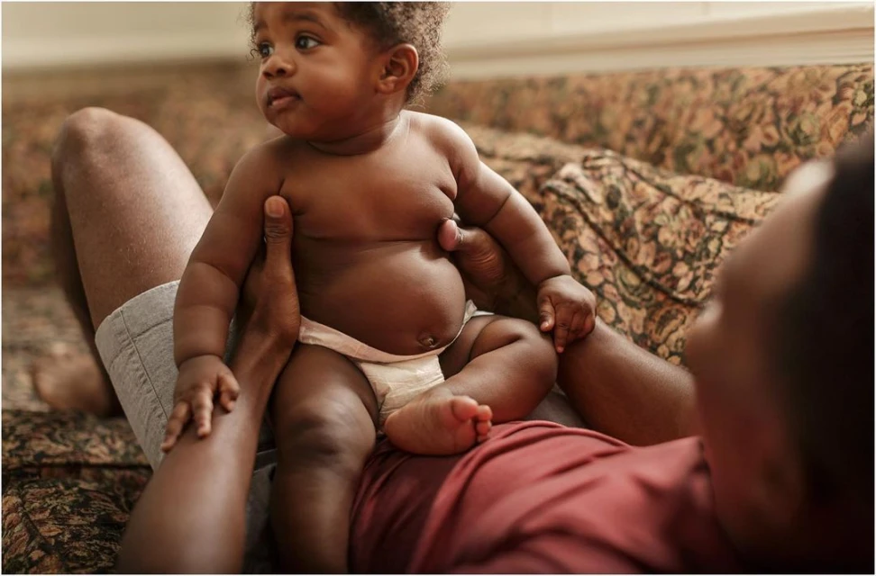 Close-up shot of a bare-chested African-American infant sitting on an adult's lap inside a home setting with a floral sofa.