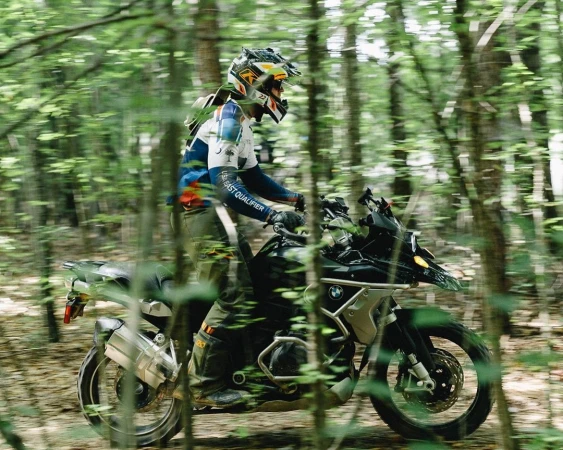 A motorcyclist in a helmet riding a black motorcycle through a dense green forest path, photographed by Shaun Fenn.