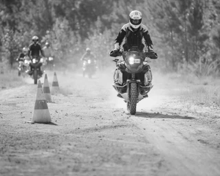 Black and white photograph of a motorcycle rider navigating a dirt course past cones with other riders behind