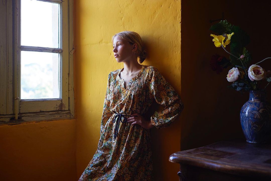 A young adult female in a floral dress stands by a window in a yellow room with a vase of flowers on a dark wooden table.