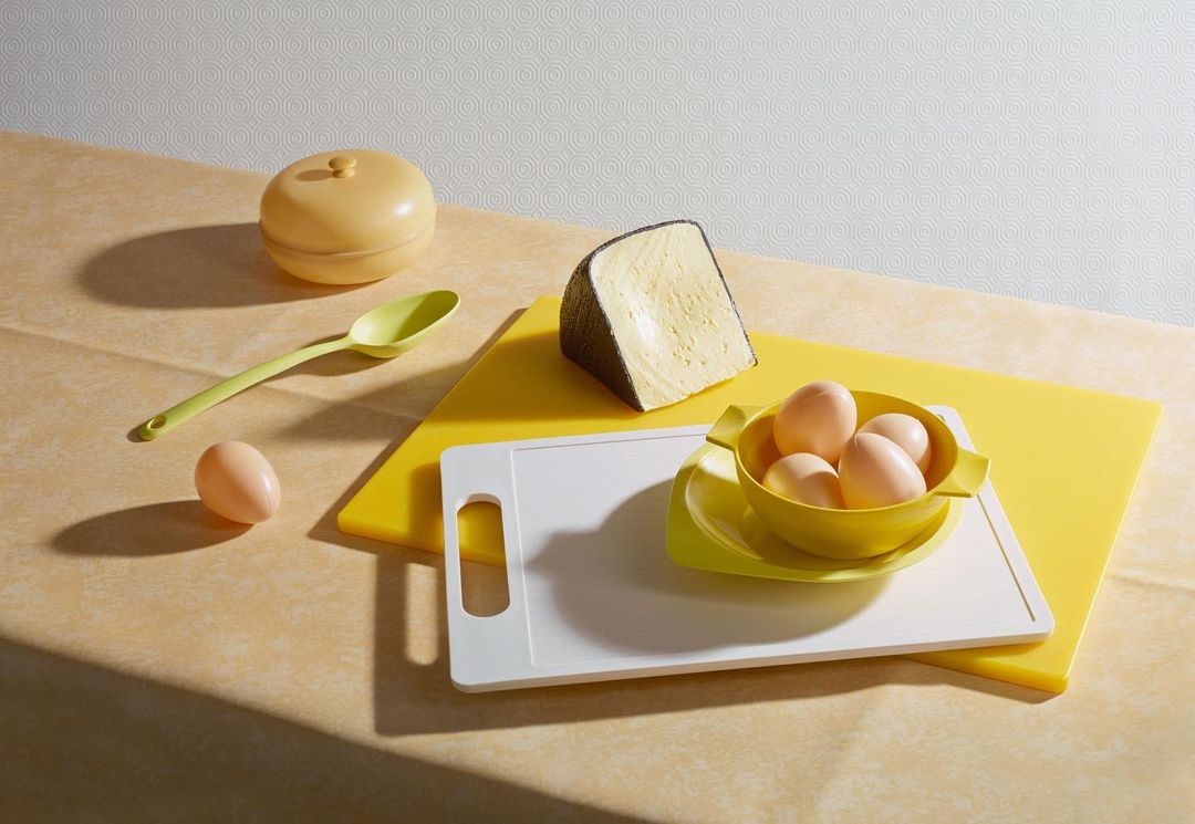 Still life featuring a cheese wedge, eggs in a yellow bowl, cutting boards, and a yellow spoon and lidded container on a patterned surface.