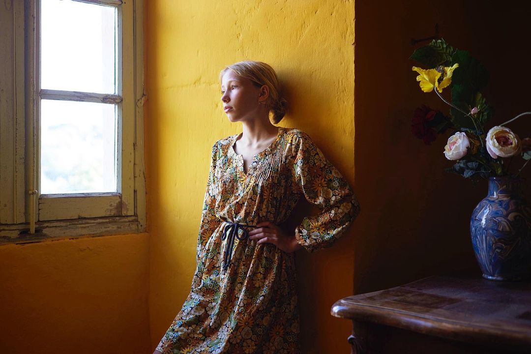 Young woman in a floral dress leans against a yellow wall next to a window, with a vase of flowers on a dark wooden table nearby.
