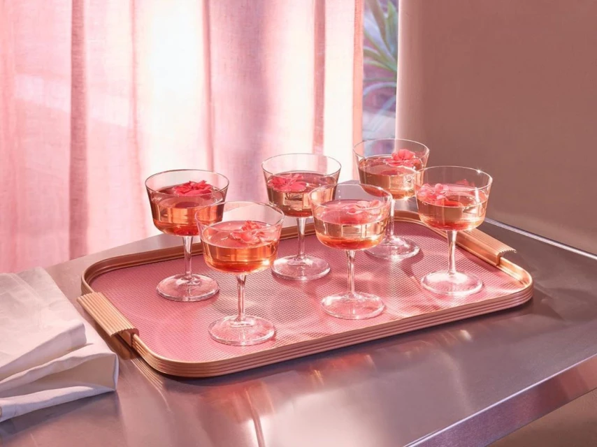 Six coupe glasses filled with pink liquid and rose petals, arranged on a pink serving tray atop a reflective surface indoors.
