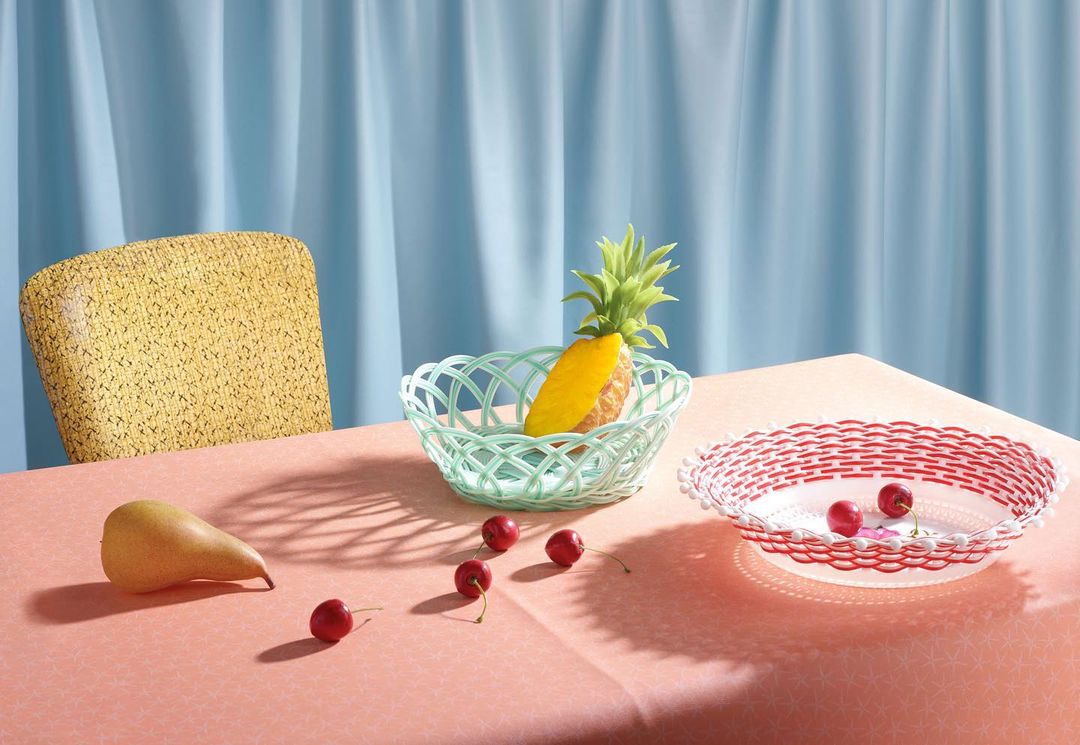 Still life with a pineapple, pear, and cherries in woven baskets on a pink tablecloth in front of a blue curtain and yellow chair.