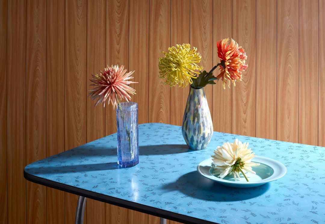 Floral arrangement featuring yellow, red, and white chrysanthemums in vases and a bowl on a blue patterned table against a wood-paneled wall.