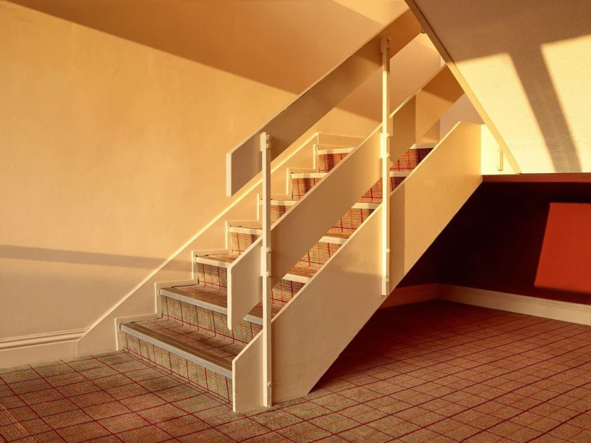 Indoor staircase with carpeted steps and landing, bathed in warm sunlight, casting shadows on the beige wall and floor.