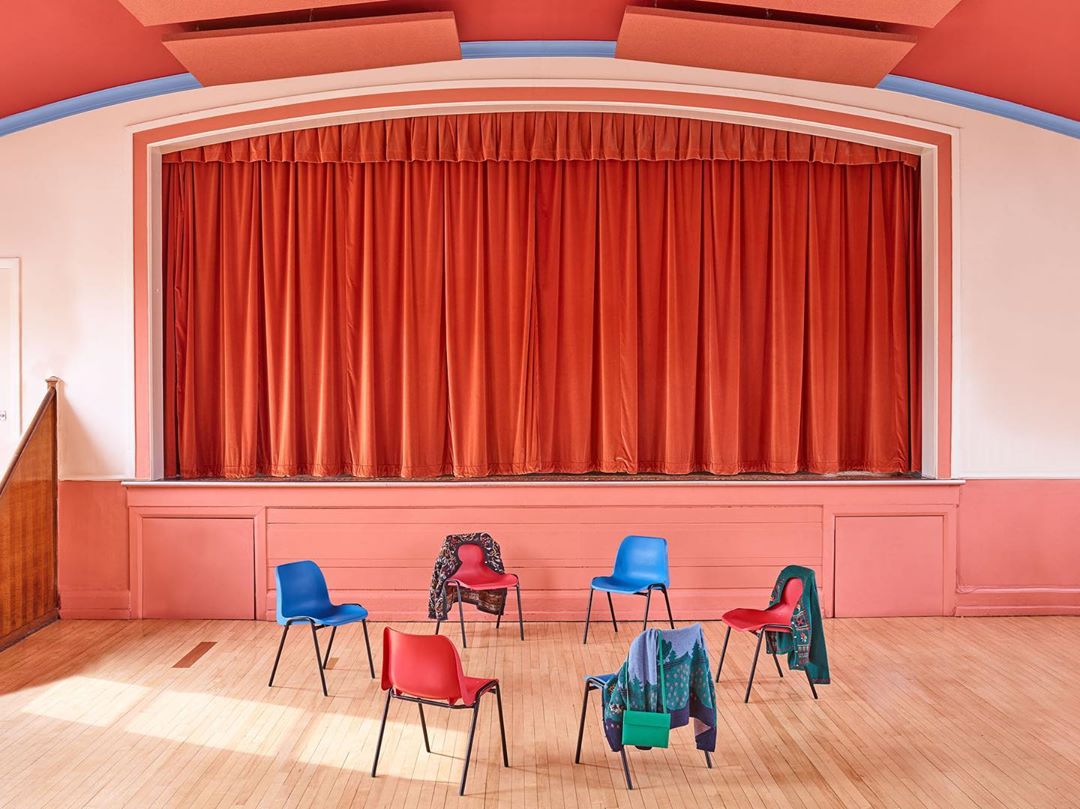 Auditorium stage with coral curtain and six colorful chairs with draped clothing in a semicircle on a light wood floor.