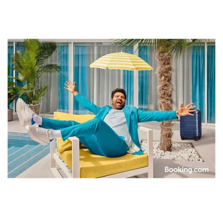 Adult male in teal suit lounges in yellow chair poolside with luggage, palm trees, and Booking.com logo visible on the right.