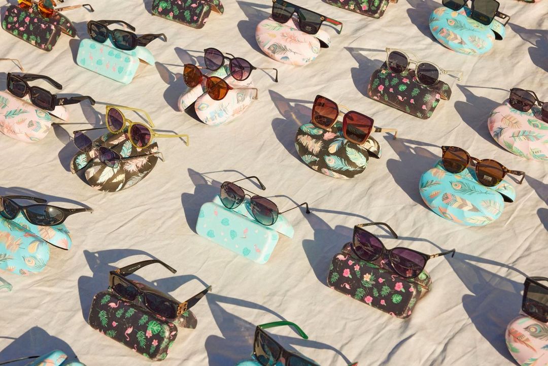 Multiple pairs of sunglasses are arranged on top of colorful patterned cases, set against a white backdrop with strong shadows.