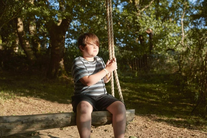 Young child in a striped t-shirt sitting on a rustic wooden rope swing in a wooded park setting.