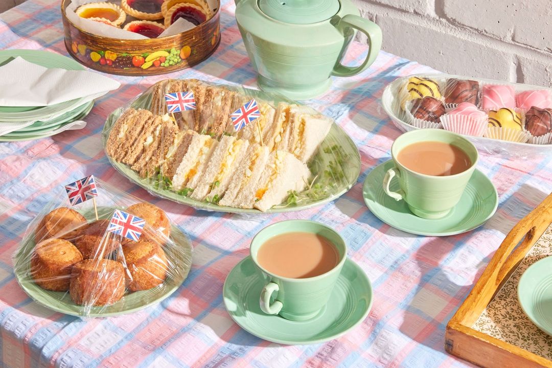 Tea party spread with sandwiches, cakes, and teacups arranged on a checkered tablecloth with sunlight casting shadows, creating a vintage feel.