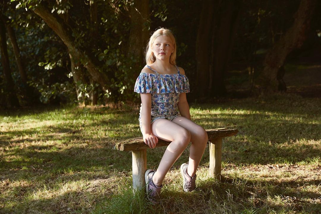 Young blonde girl seated on a rustic wooden bench in a woodland setting wearing patterned off-the-shoulder top