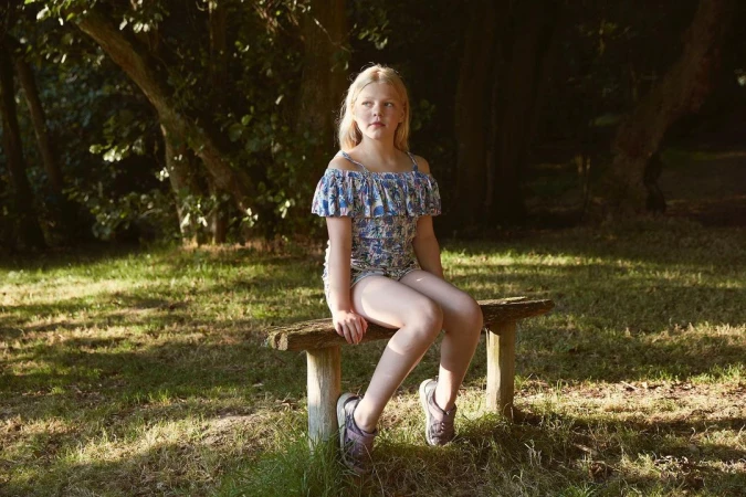 Young blonde girl seated on a rustic wooden bench in a woodland setting wearing patterned off-the-shoulder top