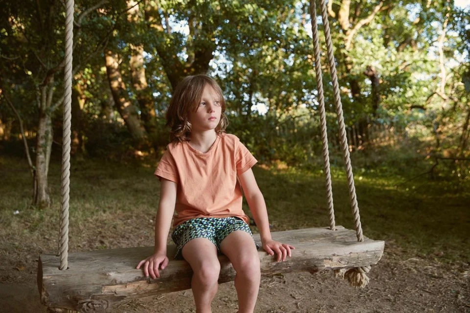 A young boy with long hair sits on a wooden tree swing in a sun-dappled outdoor woodland setting, wearing a t-shirt and patterned shorts.