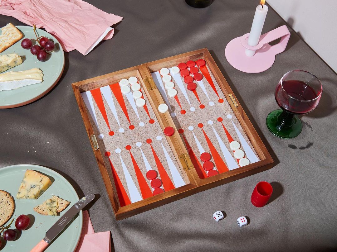 Overhead shot of an open backgammon board on a grey surface, surrounded by wine, cheese, crackers, a candle, and dice.