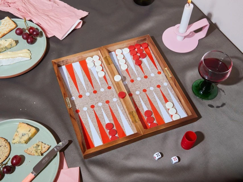 Overhead shot of an open backgammon board on a grey surface, surrounded by wine, cheese, crackers, a candle, and dice.