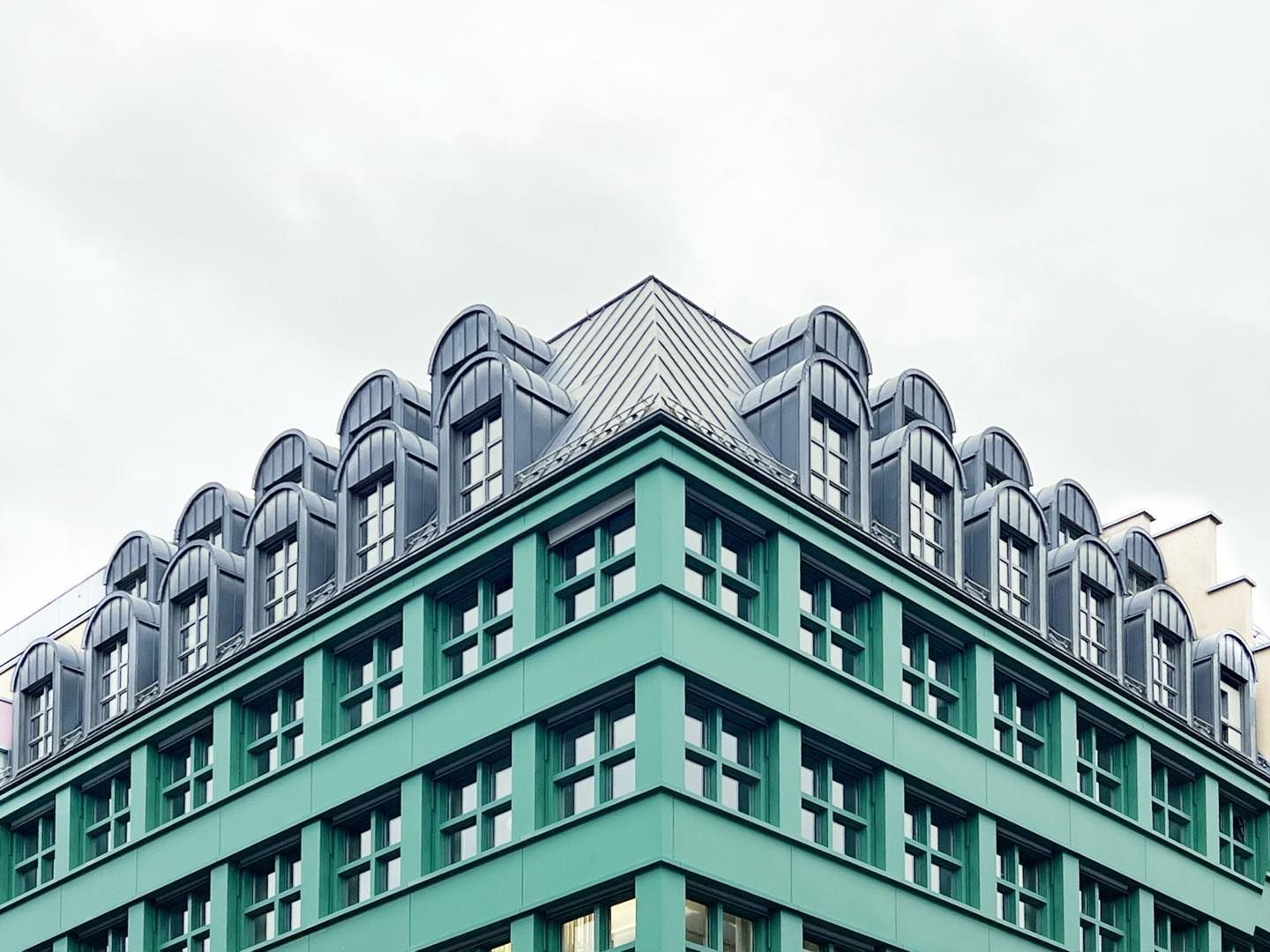 Architectural shot capturing the facade of a teal building with rows of windows and dark gray roof against a light gray sky.