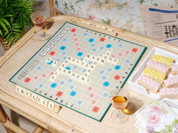 Overhead shot of a Scrabble board with letter tiles spelling words, alongside refreshments, set up on a woven rattan table.