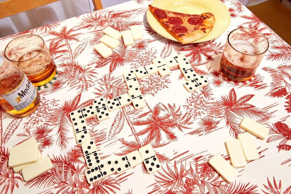 Overhead shot of dominoes on a patterned tablecloth, accompanied by a slice of pepperoni pizza, a full beer glass, and a beer bottle.