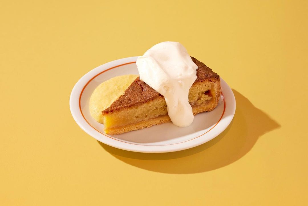 Overhead shot of a slice of cake on a plate with custard and cream, set against a vibrant yellow background with a soft shadow.