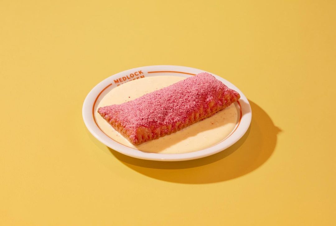Close-up of a pink-sprinkled pastry sitting on a white plate with a beige sauce, set against a bright yellow background, studio lighting.