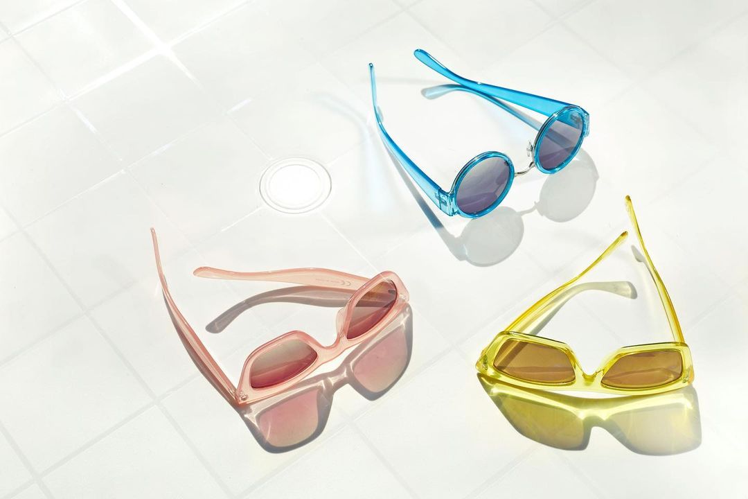 Three pairs of trendy sunglasses in pink, blue, and yellow tones are arranged on a white tiled surface with bright, diffused lighting.