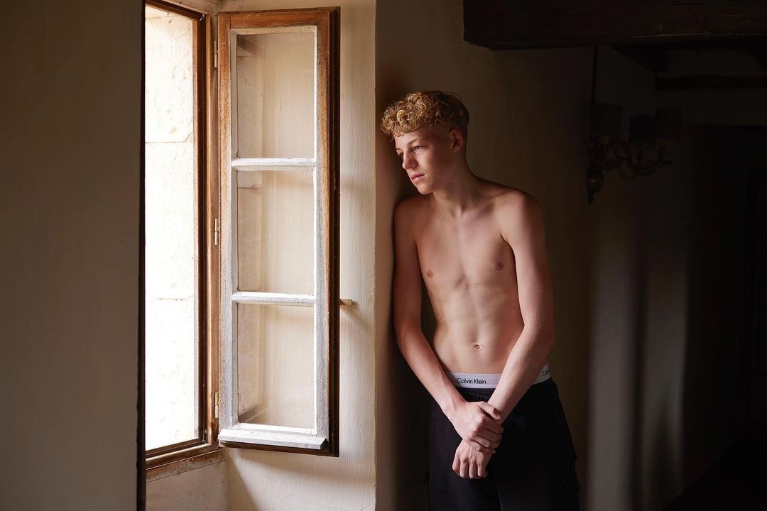 Indoor portrait of shirtless young adult male with curly blonde hair leaning against a wall near an open window in a dimly lit room.