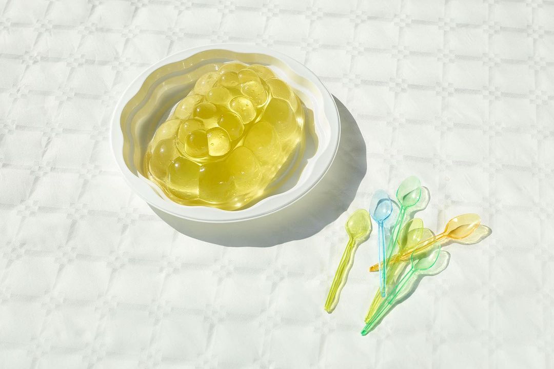 Gelatin dessert in a white bowl is accompanied by a cluster of small, colorful plastic spoons on a patterned white surface, bright sunlight.