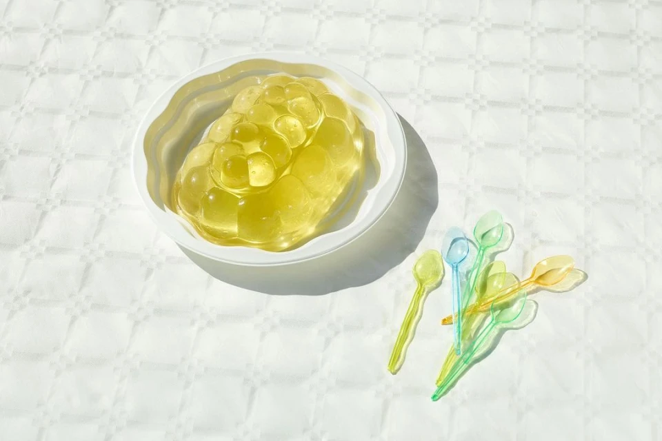 Gelatin dessert in a white bowl is accompanied by a cluster of small, colorful plastic spoons on a patterned white surface, bright sunlight.