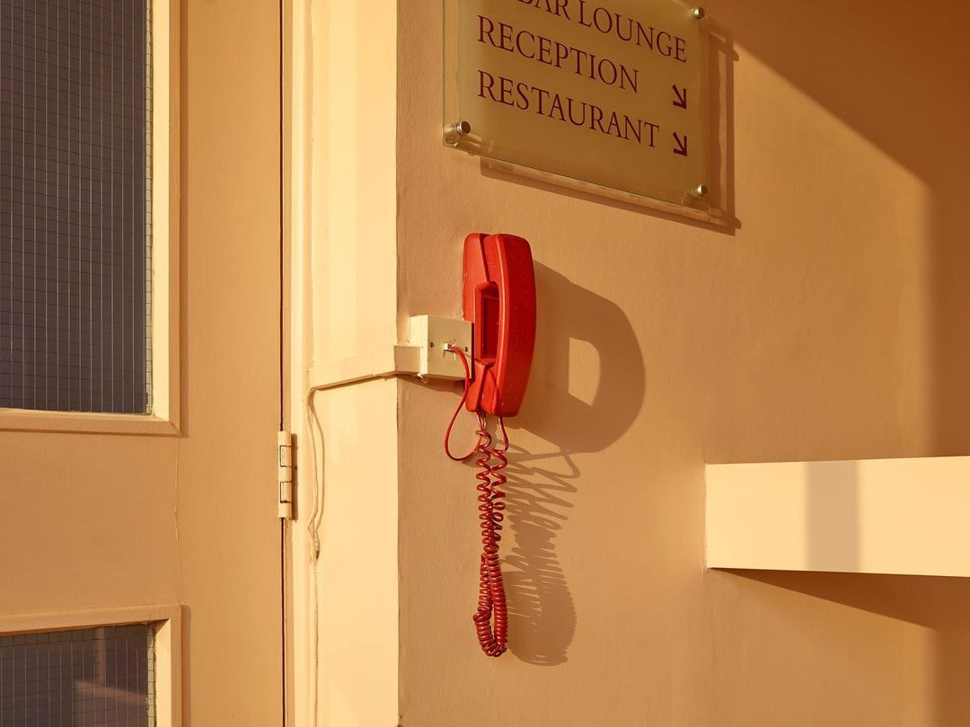 A red telephone hangs on a light beige wall next to a door with a window, illuminated by warm lighting, pointing to a reception area.