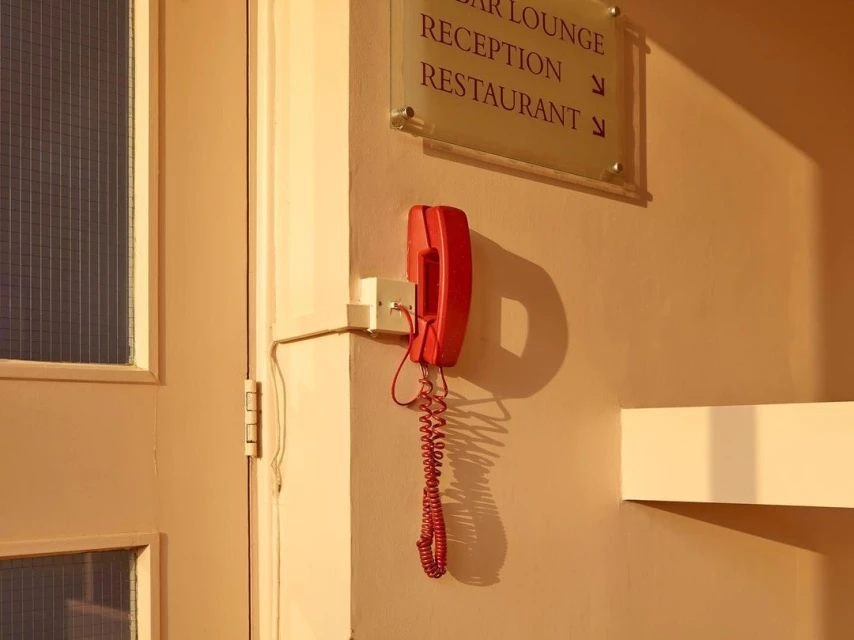 A red telephone hangs on a light beige wall next to a door with a window, illuminated by warm lighting, pointing to a reception area.
