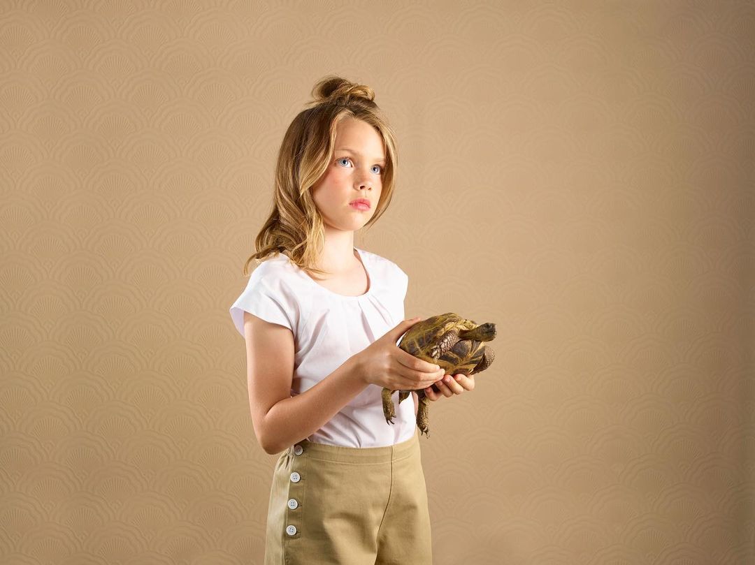 Young girl in a white shirt and tan shorts holds a turtle in a studio setting with a patterned beige background, gazing slightly upward.