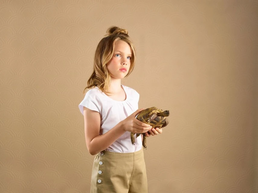 Young girl in a white shirt and tan shorts holds a turtle in a studio setting with a patterned beige background, gazing slightly upward.