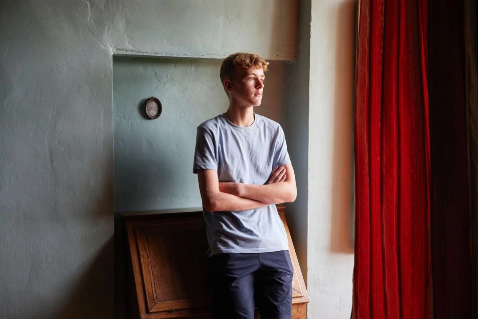 Teenage boy with blonde hair standing indoors by a window. He is wearing a blue shirt and navy pants with arms crossed.
