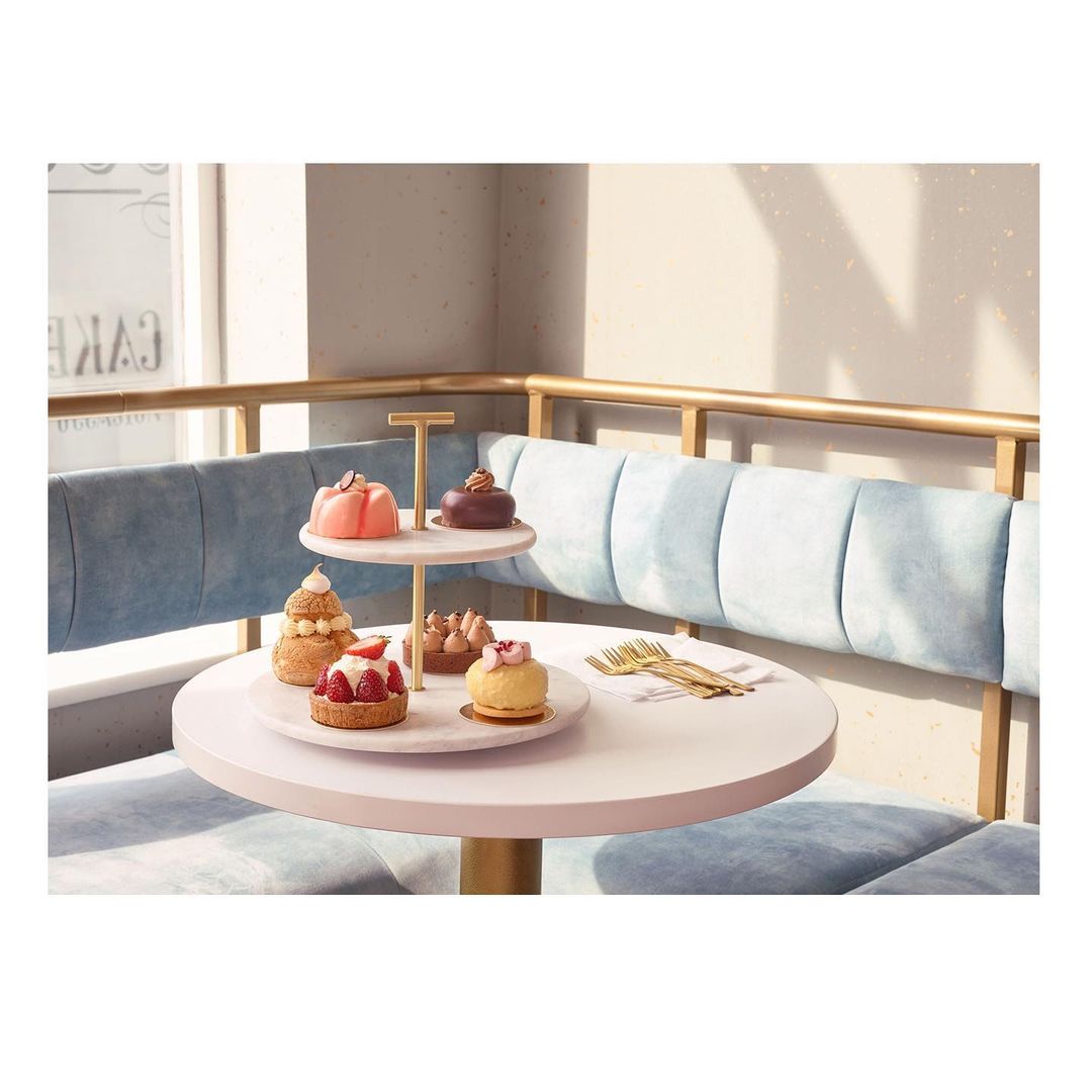 A tiered dessert stand displays assorted cakes and pastries on a table in a cozy cafe setting with natural light and blue upholstered seating.