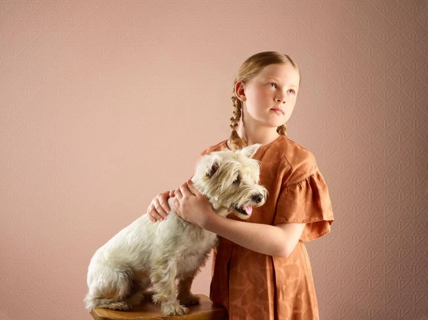 A young girl with braided hair embraces a West Highland White Terrier. The dog sits on a wooden stool against a textured peach backdrop.