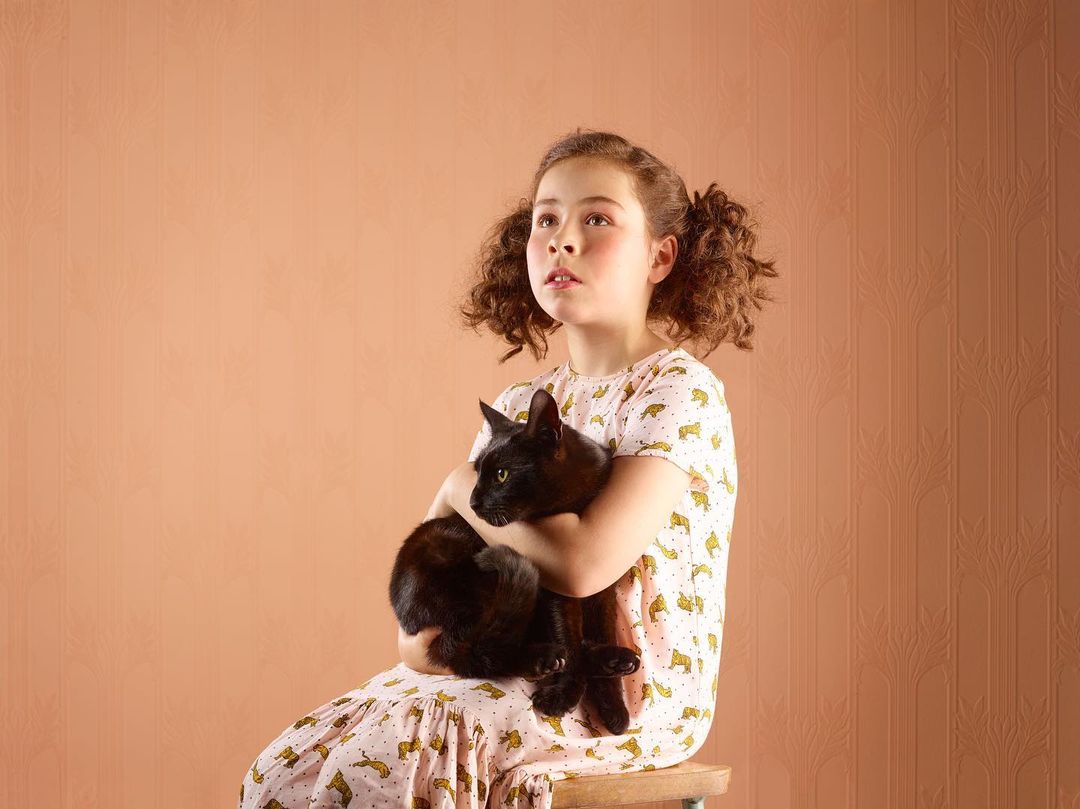 A young girl with curly pigtails is sitting on a stool, gently holding a black cat, in front of a peach colored patterned wall.