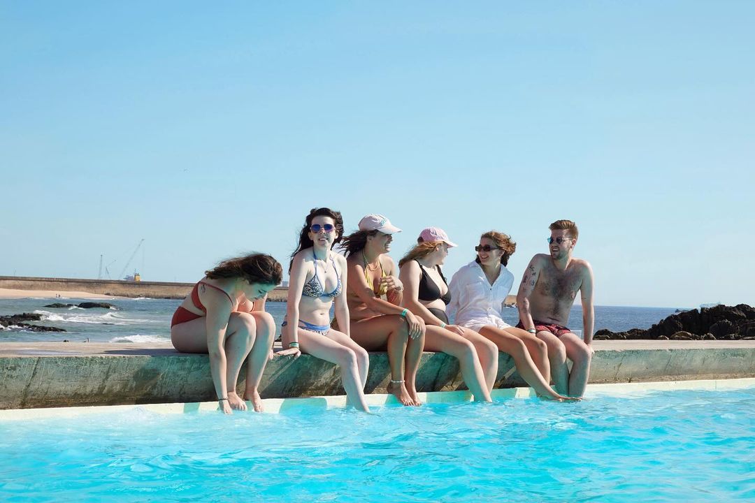 A group of six young adults sits on the edge of a pool on a sunny day, with the ocean and sky visible in the background.