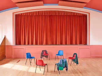 A circle of colourful chairs arranged on a wooden floor in front of a raised stage with a large red curtain.