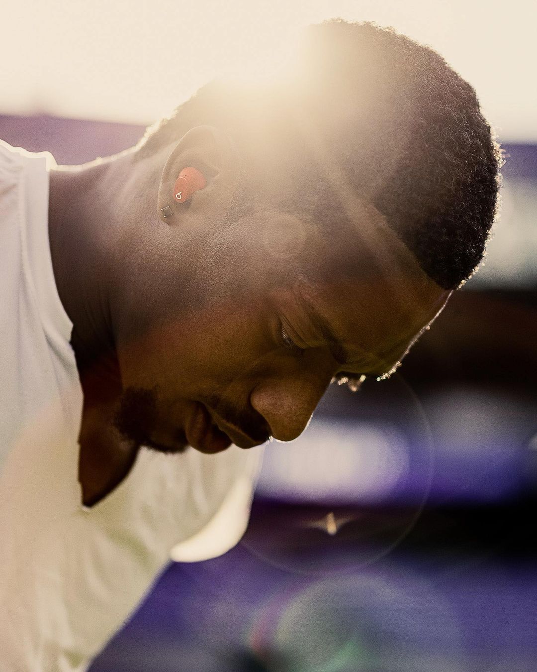 Close-up of a dark-skinned man with short hair wearing earbuds, looking down, bathed in sunlight with lens flare and purple highlights.