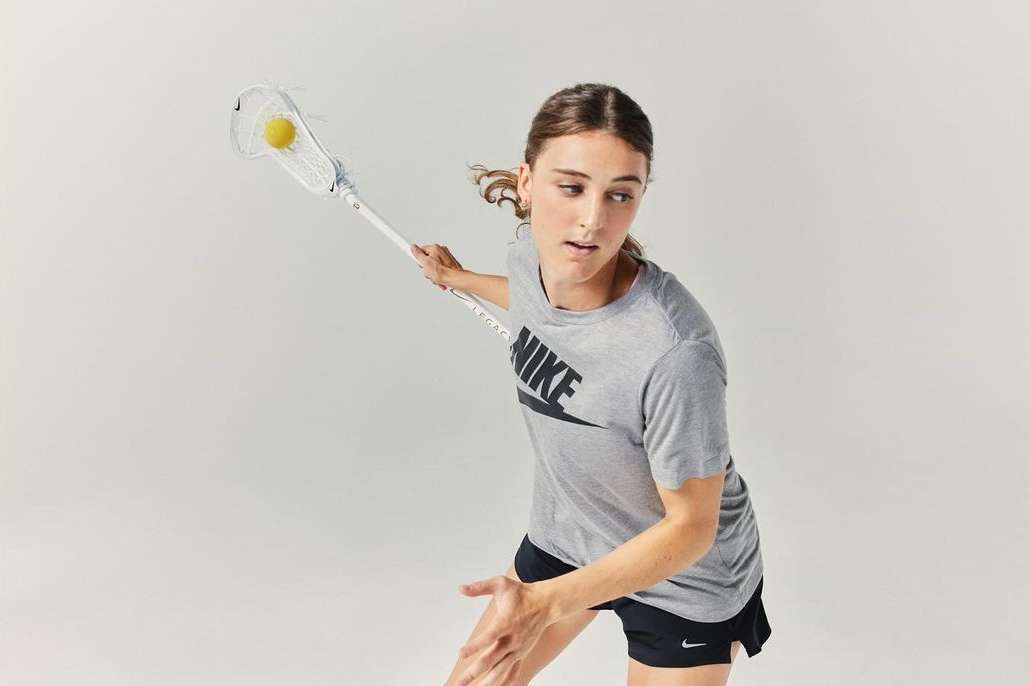 A female athlete in gray Nike shirt prepares to throw a yellow lacrosse ball with her lacrosse stick in a bright, neutral studio.