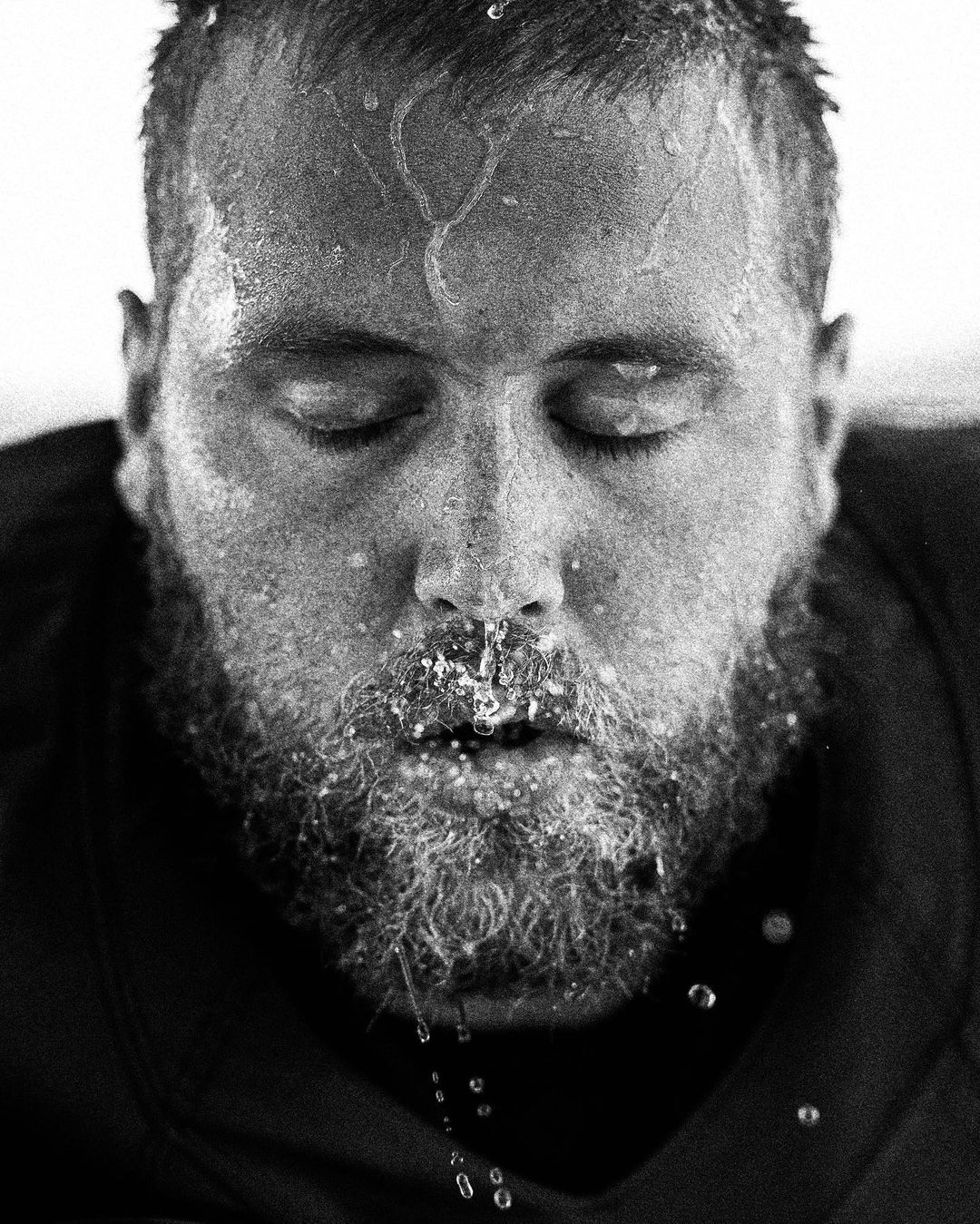 Close-up black and white portrait of a bearded adult man with eyes closed and water droplets on his face and beard.